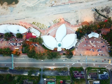 An aerial view of a uniquely designed building with three petal-shaped structures surrounded by trees and parking lots. The area appears to be under development or construction as there are large patches of bare land and construction materials visible. A major road is situated below the buildings, with cars visible on it.
