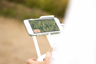 A person is holding a drone controller with a smartphone attached, displaying an aerial view of a forested area on the screen.