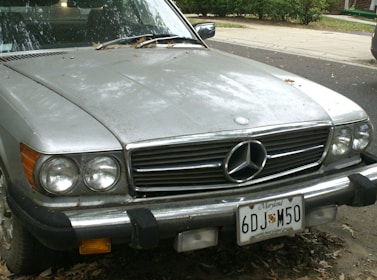 A vintage silver Mercedes-Benz car is parked on a street. There are fallen leaves on the hood, and the car has a Maryland license plate. The front headlamps and grille are visible, showing signs of age and dirt.
