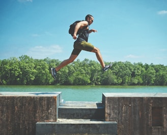 man leaping on concrete surface near body of water and forest at the distance during day