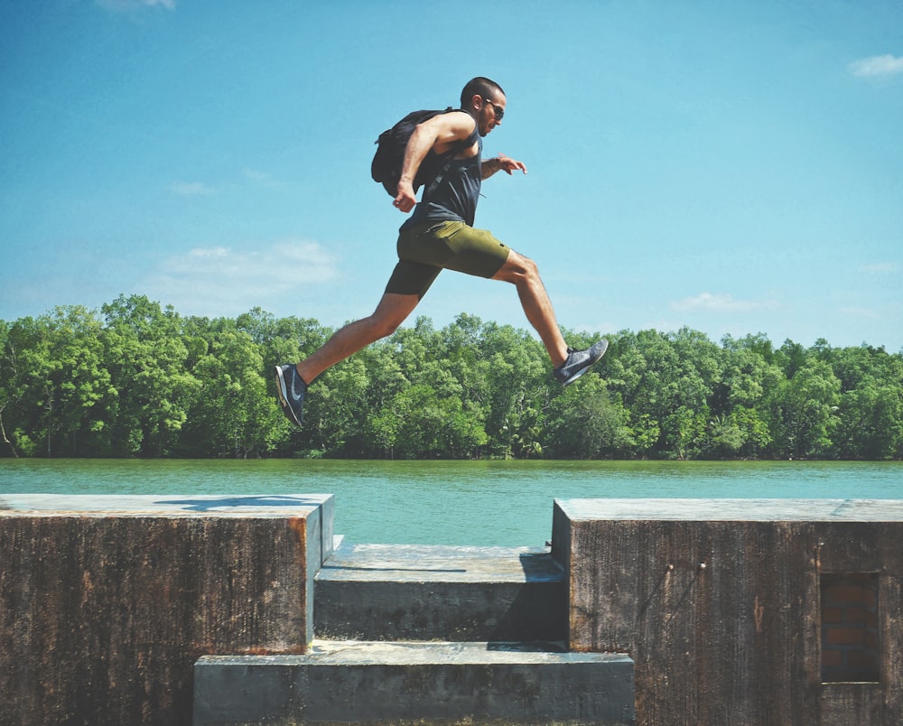 man leaping on concrete surface near body of water and forest at the distance during day