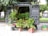 A van with open doors at the rear reveals a variety of potted plants, including small citrus trees with visible fruits and lush greenery. The scene is set outdoors on a paved area with a park and trees visible in the background. Several pots and containers are arranged on the ground in front of the van.