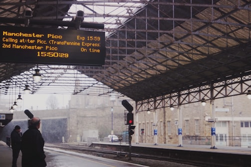 A train station platform with a large electronic sign showing information about arrivals and departures. The covered platform has lights hanging from the structure above. Two individuals are standing on the platform, looking toward the sign. The station appears relatively empty, and there is a train track running parallel to the platform.