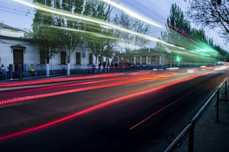 Long exposure photograph of a street at night with light trails from passing vehicles creating dynamic red and white streaks. Trees and a building are seen in the background, while several people stand on the sidewalk to the left.