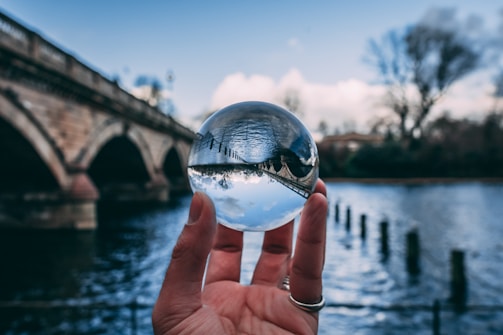 A hand holds a crystal ball with a view of a bridge and body of water reflected inside it. The background features an out-of-focus stone bridge, water, and trees under a partly cloudy sky.