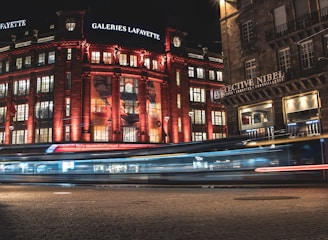 A large department store is adorned with bright red lighting, creating a dramatic contrast against the dark night sky. The building's façade features multiple large windows glowing warmly from the interior lights and has advertising banners displayed prominently. Below, a street scene is captured with the light trails of passing vehicles, suggesting a sense of movement and bustling activity.