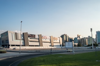 A modern urban landscape featuring a large, contemporary shopping mall with sleek architectural design, including geometric patterns on the facade. Surrounding the mall are various mid-rise buildings under a clear sky. A street with cars and green lawn is visible in the foreground.