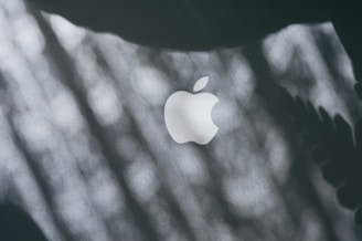 A close-up of a white Apple logo on a dark surface with dappled light and shadow patterns across the image.