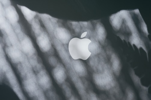 A close-up of a white Apple logo on a dark surface with dappled light and shadow patterns across the image.