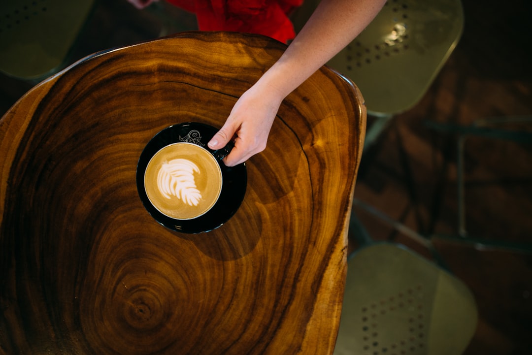 person holding teacup with cappuccino inside on top of wood slab table