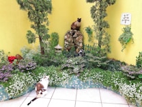 A small garden area with a yellow painted wall backdrop features lush greenery, colorful flowers, and two cats. One cat with a cream and brown coat sits perched on a rock formation, while the other, mostly white with patches of brown, sits on the tiled floor. There are small trees, purple and white flowers, and a decorative stone lantern, creating a serene and natural environment.