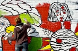 A person with short hair, wearing a dark sweater and jeans, is painting a colorful mural on a brick wall. The mural features bold designs and abstract shapes, including a large face with stylized features and vibrant colors such as red, green, yellow, and black. The artistic style is bold and graphic, with thick black outlines and bright colors filling in the shapes.