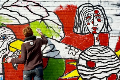 A person with short hair, wearing a dark sweater and jeans, is painting a colorful mural on a brick wall. The mural features bold designs and abstract shapes, including a large face with stylized features and vibrant colors such as red, green, yellow, and black. The artistic style is bold and graphic, with thick black outlines and bright colors filling in the shapes.