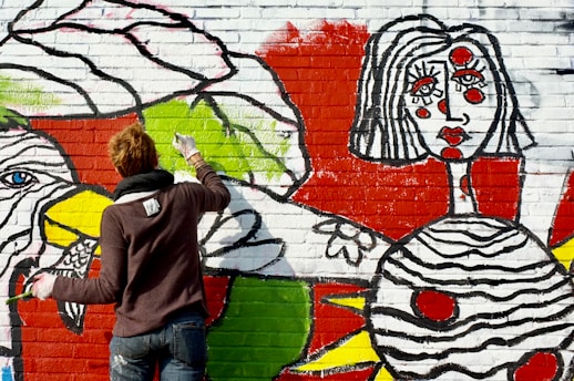 A person with short hair, wearing a dark sweater and jeans, is painting a colorful mural on a brick wall. The mural features bold designs and abstract shapes, including a large face with stylized features and vibrant colors such as red, green, yellow, and black. The artistic style is bold and graphic, with thick black outlines and bright colors filling in the shapes.