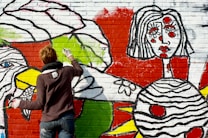 A person with short hair, wearing a dark sweater and jeans, is painting a colorful mural on a brick wall. The mural features bold designs and abstract shapes, including a large face with stylized features and vibrant colors such as red, green, yellow, and black. The artistic style is bold and graphic, with thick black outlines and bright colors filling in the shapes.