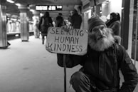 A bearded man in a subway station is holding a sign that reads 'Seeking Human Kindness'. He is wearing a beanie and a jacket, sitting on the ground with a somber expression. People are walking in the background, appearing somewhat blurred, suggesting movement and a bustling environment.