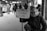 A bearded man in a subway station is holding a sign that reads 'Seeking Human Kindness'. He is wearing a beanie and a jacket, sitting on the ground with a somber expression. People are walking in the background, appearing somewhat blurred, suggesting movement and a bustling environment.