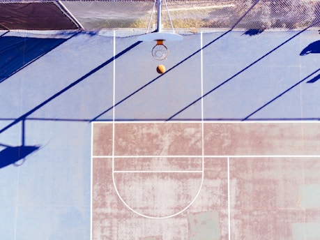 An aerial view of a basketball court with a basketball in mid-air approaching the hoop. The court is marked with white lines and has a worn, faded appearance. Shadows from a fence are cast across the court, creating a geometric pattern.