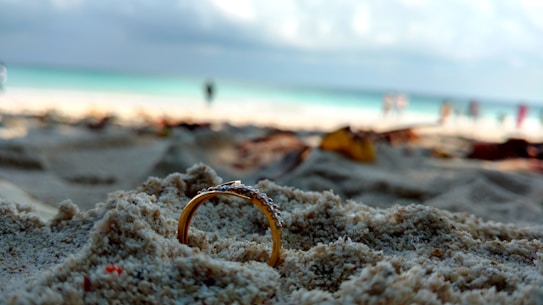 A gold ring partially buried in sandy beach, with a blurred background including the ocean and a few distant figures.