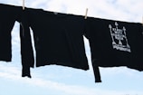 Black long-sleeve shirts are hanging on a clothesline against a bright, partly cloudy sky. One shirt features a design with arrows and the phrase 'Stand With Standing Rock'.