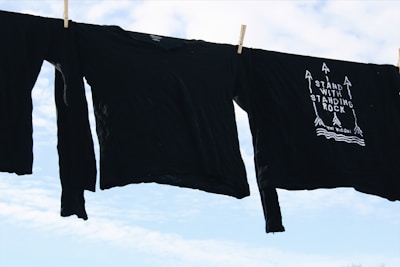 Black long-sleeve shirts are hanging on a clothesline against a bright, partly cloudy sky. One shirt features a design with arrows and the phrase 'Stand With Standing Rock'.