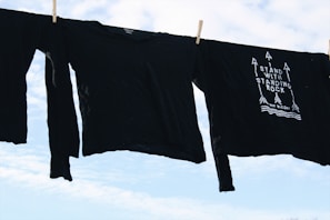 Black long-sleeve shirts are hanging on a clothesline against a bright, partly cloudy sky. One shirt features a design with arrows and the phrase 'Stand With Standing Rock'.