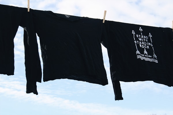 Black long-sleeve shirts are hanging on a clothesline against a bright, partly cloudy sky. One shirt features a design with arrows and the phrase 'Stand With Standing Rock'.