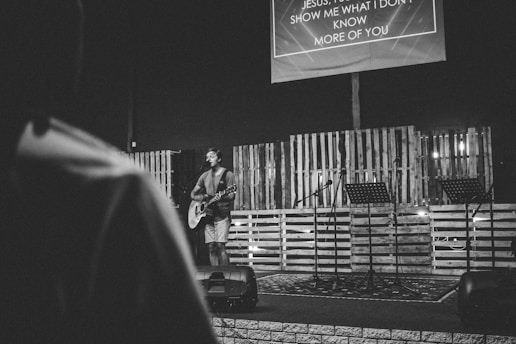 A person with a guitar stands on a stage surrounded by wooden pallets, creating a rustic backdrop. The stage features several microphones and music stands. Above, a large screen displays lyrics. The setting suggests an informal musical performance or worship event in a darkened room.