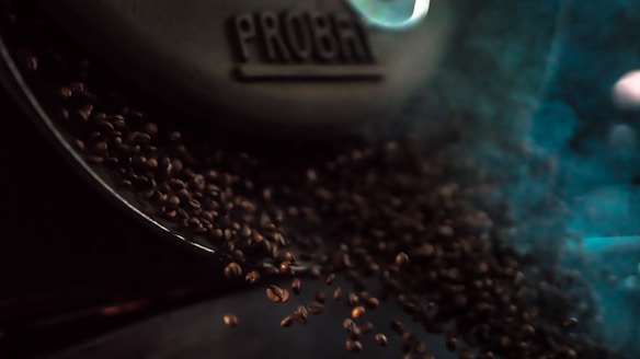 Coffee beans are being roasted, tumbling out of a machine labeled 'PROBAT' amidst a cloud of steam or smoke.