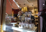 A man inside a shop is arranging chocolate-covered strawberries in paper cones on a tiered stand. The shop's interior is warmly lit, with multiple reflections visible on the glass window, adding to the cozy atmosphere of the confectionery display. Outside, traces of a busy street and colorful umbrellas are faintly visible.