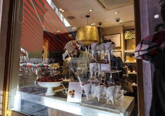 A man inside a shop is arranging chocolate-covered strawberries in paper cones on a tiered stand. The shop's interior is warmly lit, with multiple reflections visible on the glass window, adding to the cozy atmosphere of the confectionery display. Outside, traces of a busy street and colorful umbrellas are faintly visible.