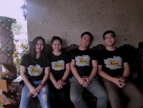 Four people are sitting side by side on a wooden bench. They are wearing matching black T-shirts with the words 'Team Building' printed prominently on the front. The background features a textured stone wall and some decorative plants. One person is wearing a watch, and the overall composition suggests a relaxed, friendly atmosphere.