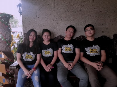 Four people are sitting side by side on a wooden bench. They are wearing matching black T-shirts with the words 'Team Building' printed prominently on the front. The background features a textured stone wall and some decorative plants. One person is wearing a watch, and the overall composition suggests a relaxed, friendly atmosphere.