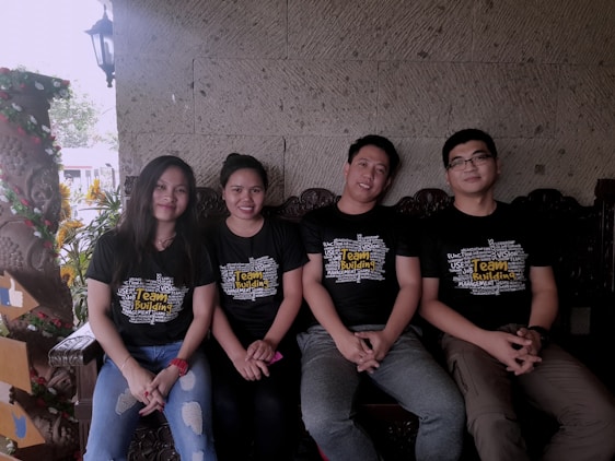 Four people are sitting side by side on a wooden bench. They are wearing matching black T-shirts with the words 'Team Building' printed prominently on the front. The background features a textured stone wall and some decorative plants. One person is wearing a watch, and the overall composition suggests a relaxed, friendly atmosphere.