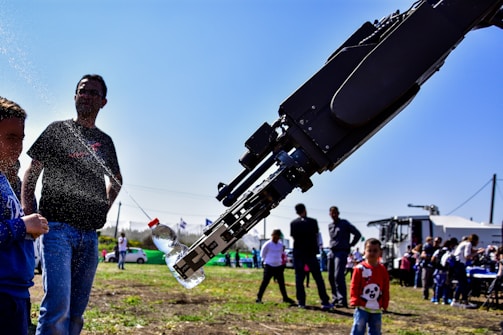 A robotic arm pours a stream of liquid from a bottle, surrounded by several people including children. The setting is outdoors on a sunny day, with a clear blue sky and groups of people visible in the background, presumably at an event or exhibition. The ground is grassy, and some vehicles or equipment are seen in the background.