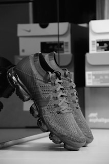 A pair of knitted sneakers with a sleek design featuring a transparent air cushioning sole. The sneakers are positioned against a reflective surface, and several shoeboxes are faintly visible in the background.