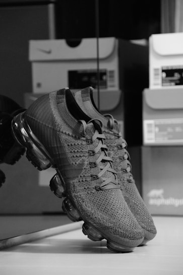 A pair of knitted sneakers with a sleek design featuring a transparent air cushioning sole. The sneakers are positioned against a reflective surface, and several shoeboxes are faintly visible in the background.