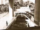 A group of people stands on an elevated walkway overlooking an archaeological site. The setting includes large concrete columns and structures, with trees and stairs visible in the background.