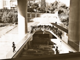 A group of people stands on an elevated walkway overlooking an archaeological site. The setting includes large concrete columns and structures, with trees and stairs visible in the background.