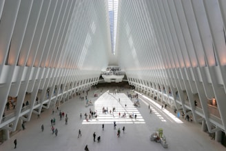 A spacious and modern architectural space with a large open floor and high, white rib-like structures forming an arched ceiling. Sunlight streams in through the top, creating patterns of light and shadow on the ground. People of various ages are walking or standing throughout the open area, adding a sense of liveliness to the scene.