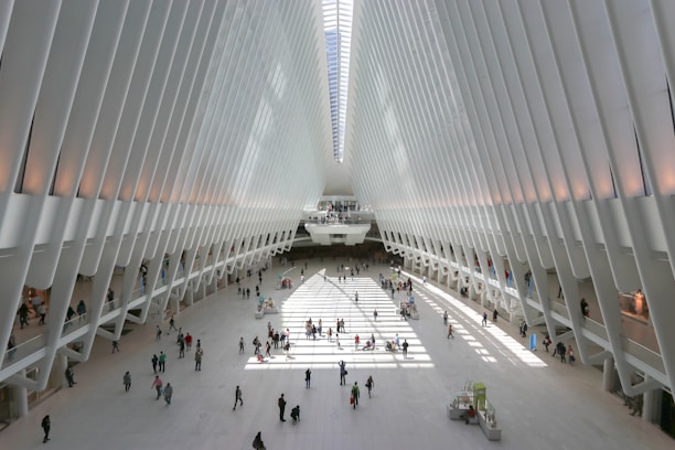 A spacious and modern architectural space with a large open floor and high, white rib-like structures forming an arched ceiling. Sunlight streams in through the top, creating patterns of light and shadow on the ground. People of various ages are walking or standing throughout the open area, adding a sense of liveliness to the scene.