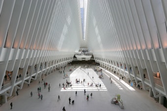 A spacious and modern architectural space with a large open floor and high, white rib-like structures forming an arched ceiling. Sunlight streams in through the top, creating patterns of light and shadow on the ground. People of various ages are walking or standing throughout the open area, adding a sense of liveliness to the scene.
