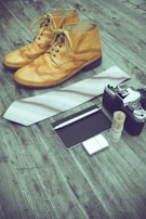 A pair of vintage-style brown leather shoes is placed on a rustic wooden surface alongside a white tie with subtle stripes. Nearby, there is an old-fashioned camera and a rolled-up stack of bills secured with a rubber band. A small notepad with a pen rests beside a metal lighter.