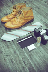 A pair of vintage-style brown leather shoes is placed on a rustic wooden surface alongside a white tie with subtle stripes. Nearby, there is an old-fashioned camera and a rolled-up stack of bills secured with a rubber band. A small notepad with a pen rests beside a metal lighter.