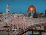 A historic city landscape featuring a prominent building with a large, golden dome. The foreground showcases ancient stone structures and a bustling area with people milling about. A tall tower with a blue dome is visible, adding to the skyline of the surroundings.