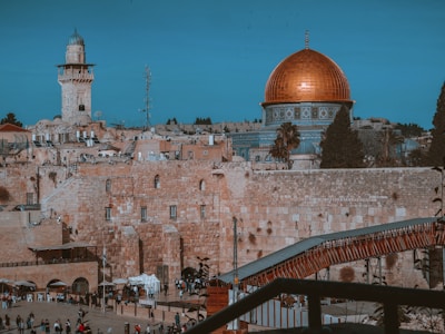A historic city landscape featuring a prominent building with a large, golden dome. The foreground showcases ancient stone structures and a bustling area with people milling about. A tall tower with a blue dome is visible, adding to the skyline of the surroundings.