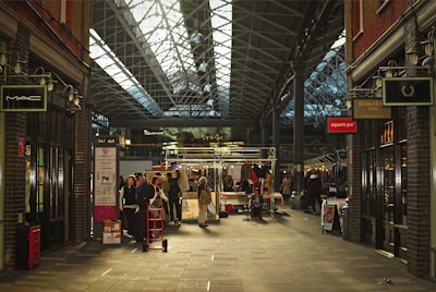 A bustling indoor market with a high, glass-paneled roof and exposed metal framework. Shoppers and vendors gather around stalls displaying various items. The atmosphere is lively, with people walking and exploring the merchandise. Brick walls and storefronts line both sides of the walkway, featuring signs from various shops.