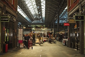 A bustling indoor market with a high, glass-paneled roof and exposed metal framework. Shoppers and vendors gather around stalls displaying various items. The atmosphere is lively, with people walking and exploring the merchandise. Brick walls and storefronts line both sides of the walkway, featuring signs from various shops.