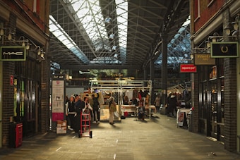 A bustling indoor market with a high, glass-paneled roof and exposed metal framework. Shoppers and vendors gather around stalls displaying various items. The atmosphere is lively, with people walking and exploring the merchandise. Brick walls and storefronts line both sides of the walkway, featuring signs from various shops.