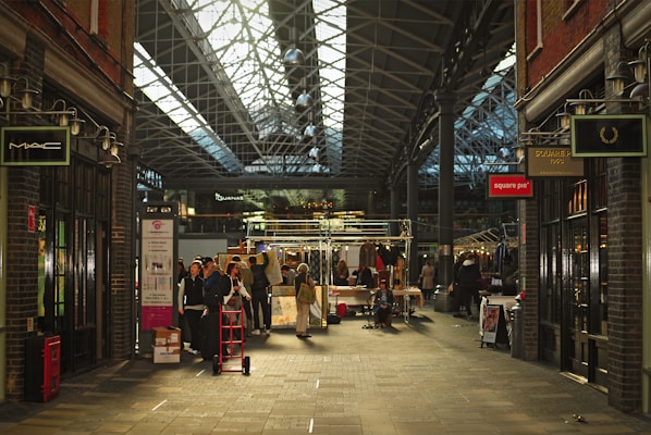 A bustling indoor market with a high, glass-paneled roof and exposed metal framework. Shoppers and vendors gather around stalls displaying various items. The atmosphere is lively, with people walking and exploring the merchandise. Brick walls and storefronts line both sides of the walkway, featuring signs from various shops.